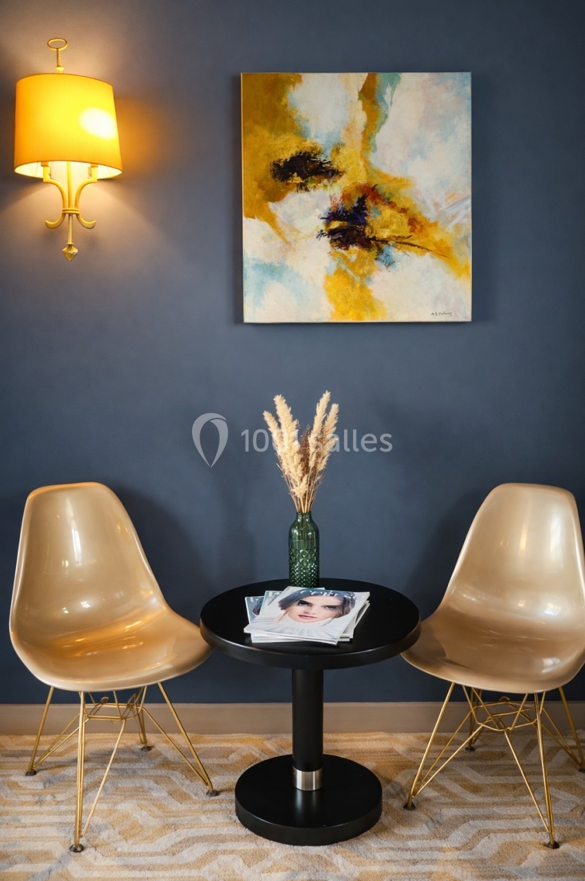Deux chaises beige entourent une table noire avec un vase de pampas et des magazines, sur fond de mur bleu.