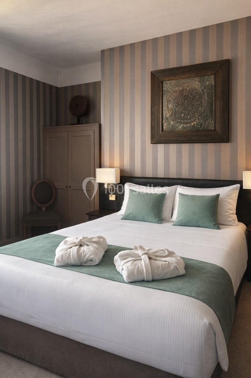 Chambre avec lit double, draps blancs, coussins verts, peignoirs pliés, mur à rayures et éclairage doux.