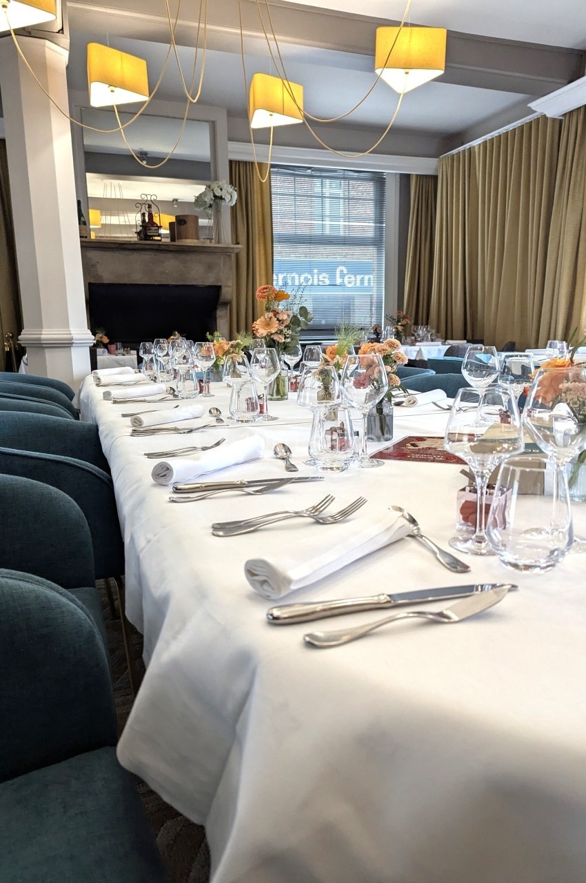 Table dressée avec nappes blanches, couverts, verres et décorations florales dans une salle lumineuse avec grandes fenêtres.