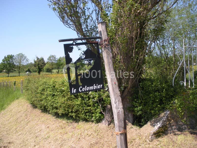 Panneau en métal noir ’Le Colombier’ suspendu à un poteau en bois, devant une haie et un paysage champêtre.