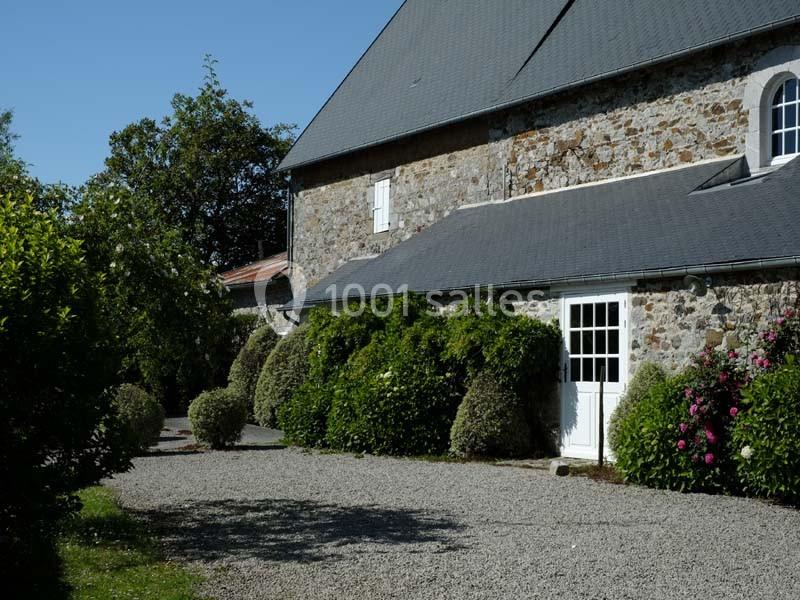 Cour gravillonnée devant une maison en pierre avec des arbustes taillés et des fleurs sous un ciel dégagé.