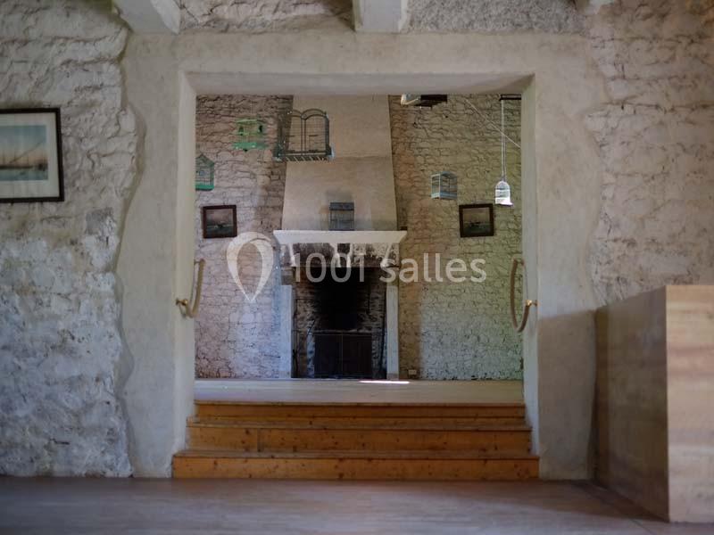 Vue d'une pièce en pierre avec une cheminée centrale, des murs décorés de cadres et un plafond en poutres apparentes.