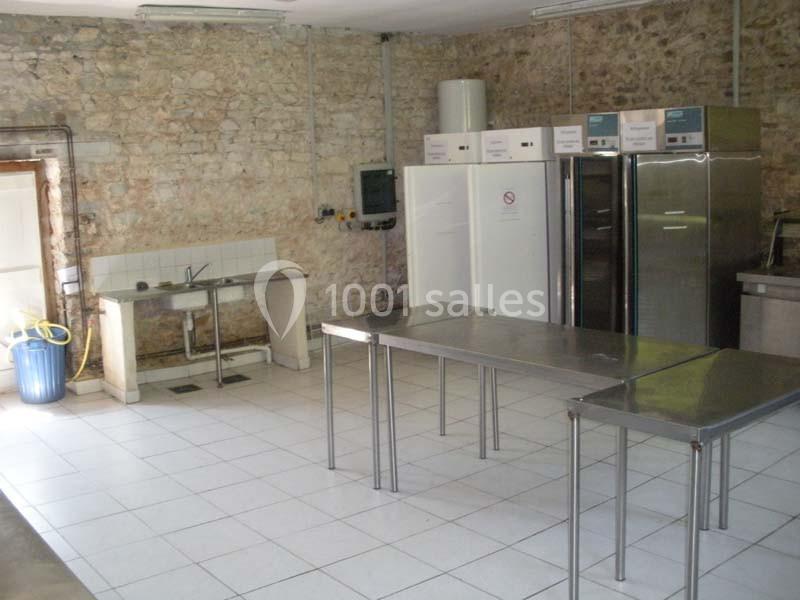 Cuisine professionnelle avec murs en pierre, plans de travail en inox, éviers, réfrigérateurs et carrelage blanc au sol.