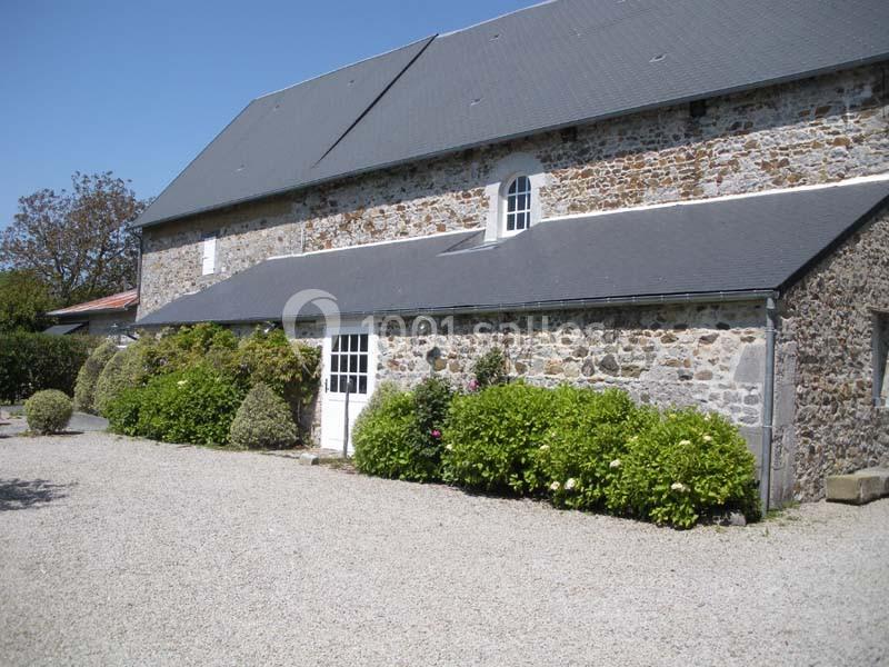 Façade en pierre d'une maison avec toit en ardoise, porte blanche, buissons et allée gravillonnée.