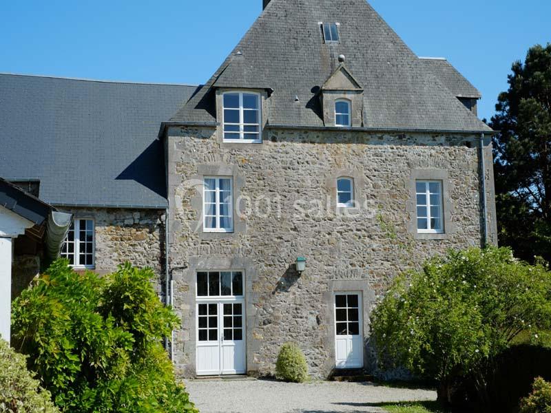 Façade en pierre d'une maison traditionnelle avec toit en ardoise, entourée de verdure sous un ciel bleu.