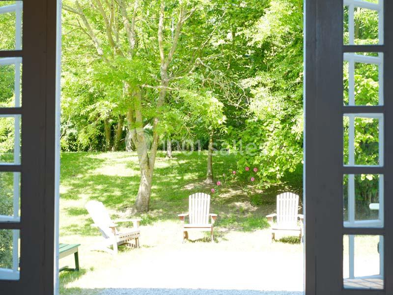 Vue depuis une porte ouverte sur un jardin verdoyant avec des arbres et des chaises en bois disposées sur la pelouse.