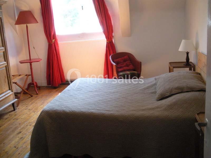 Chambre avec lit double, parquet en bois, rideaux rouges, lampe de chevet et fauteuil près d'une fenêtre.