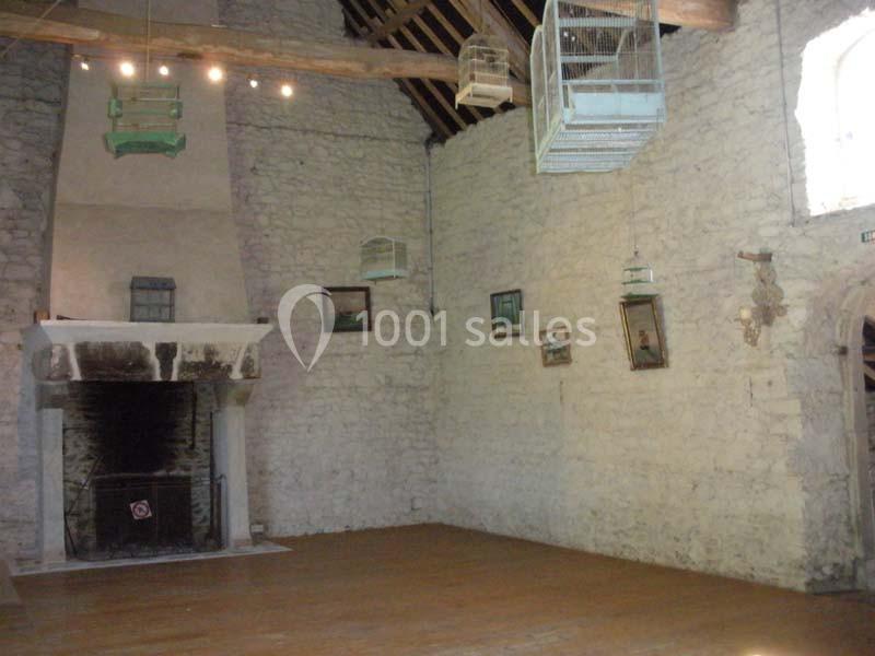 Salle en pierre avec cheminée ancienne, plafond en poutres apparentes et cages suspendues, décorée de tableaux.