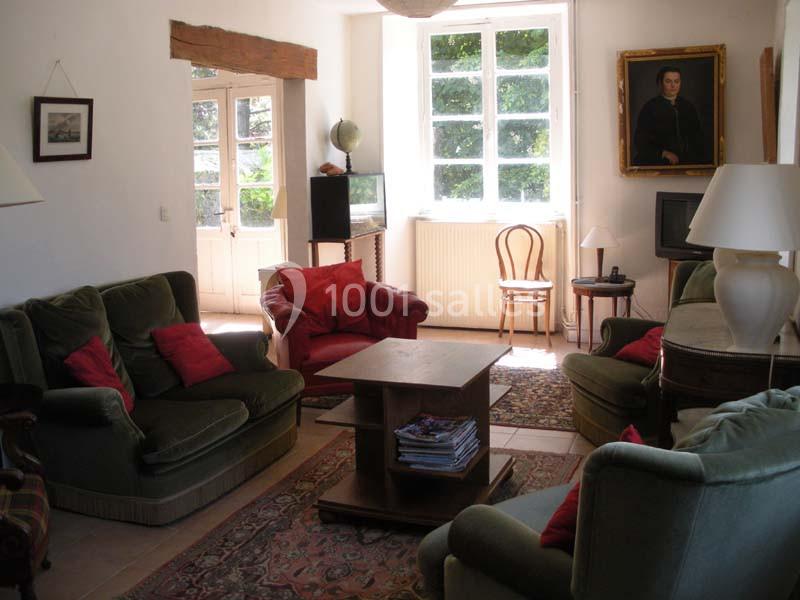 Salon lumineux avec canapés verts, coussins rouges, table basse en bois, tapis, et grandes fenêtres donnant sur un jardin.