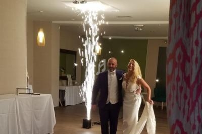 Un couple en tenue de mariage entre dans une salle éclairée, accompagné d'effets pyrotechniques en arrière-plan.