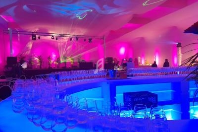 Salle de réception illuminée de lumières roses et bleues, avec un bar central et des verres disposés.