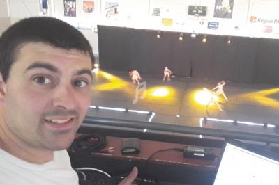 Homme souriant en premier plan dans une salle de spectacle, avec une scène éclairée et des danseurs en arrière-plan.