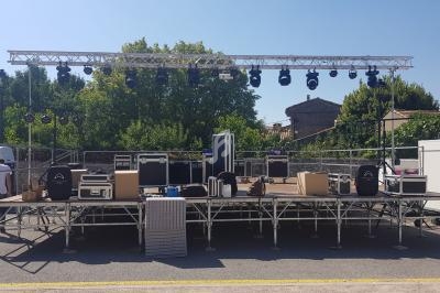 Scène extérieure en cours de montage avec projecteurs, enceintes et matériel technique disposés sur l'estrade.