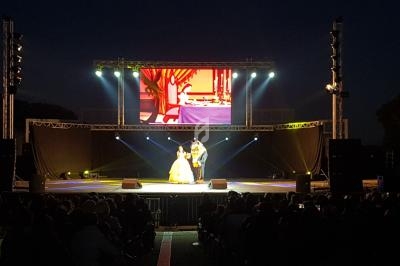 Un spectacle en plein air avec deux artistes sur scène éclairée, devant un public assis, et un écran géant en arrière-plan.