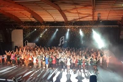 Un grand groupe de personnes sur scène dans une salle, levant les bras sous des projecteurs et des confettis.