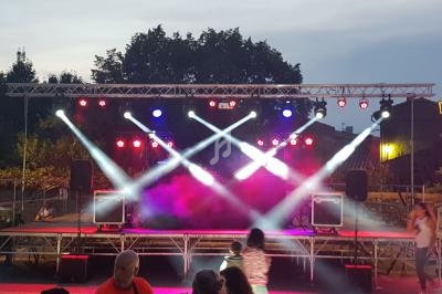 Scène extérieure éclairée par des projecteurs colorés, avec quelques personnes au premier plan en soirée.