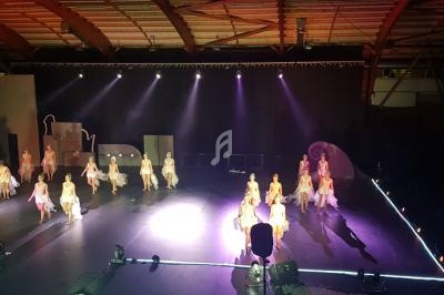 Danseuses en costume sur une scène éclairée, dans une salle avec une structure en bois visible en arrière-plan.