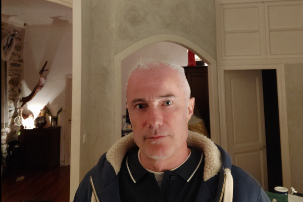 Homme aux cheveux courts blancs dans une pièce avec des murs clairs, une arche et des meubles en arrière-plan.