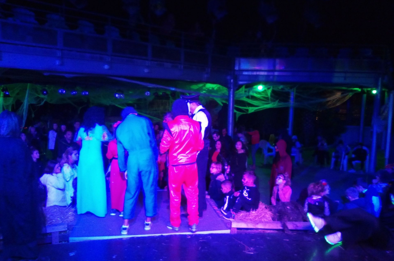 Groupe de personnes déguisées sur une scène éclairée en bleu et vert, entourées de spectateurs assis.