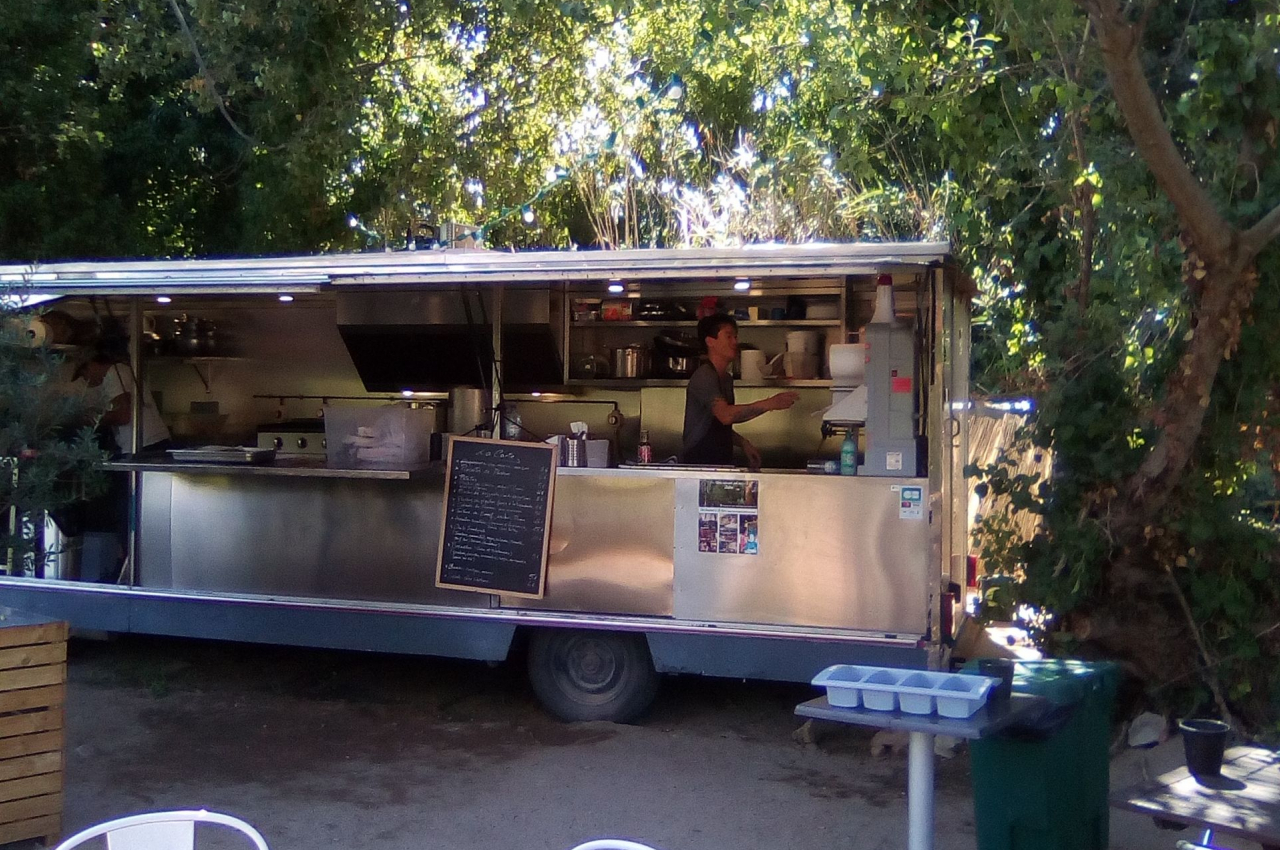 Camion de restauration mobile en plein air, entouré d'arbres, avec une table et des chaises au premier plan.