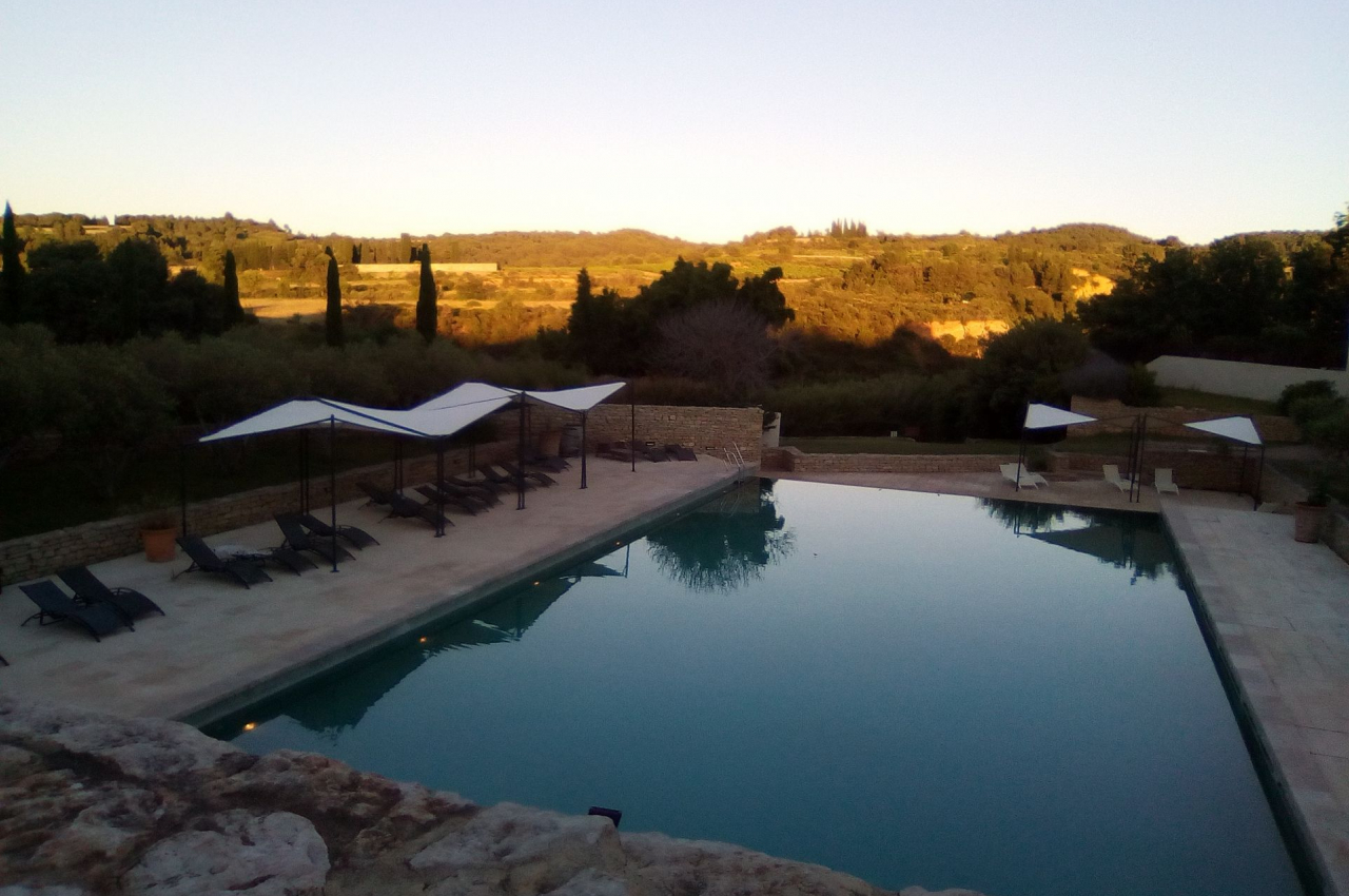 Piscine extérieure entourée de chaises longues et parasols, avec vue sur un paysage vallonné au coucher du soleil.