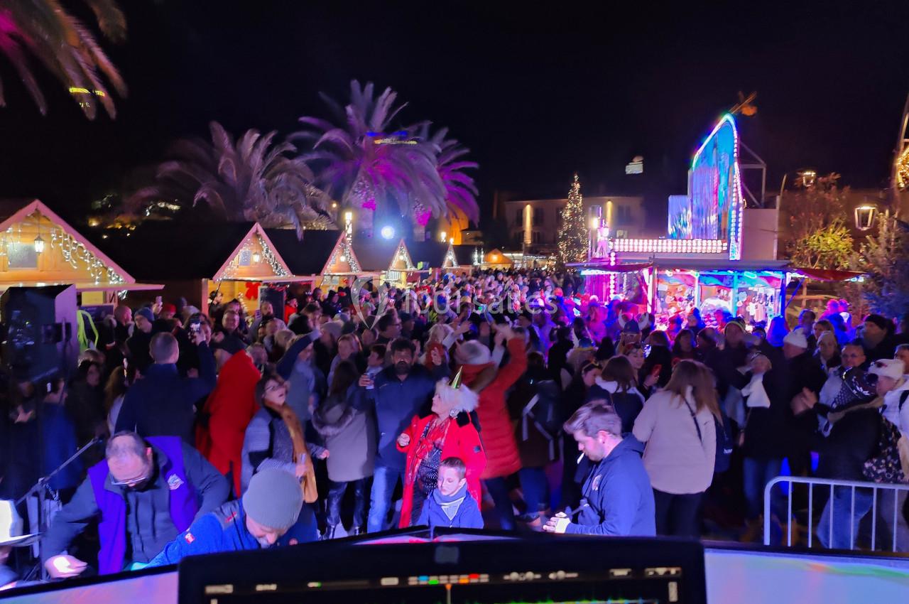 Foule rassemblée de nuit dans un marché illuminé, avec chalets décorés et palmiers en arrière-plan.