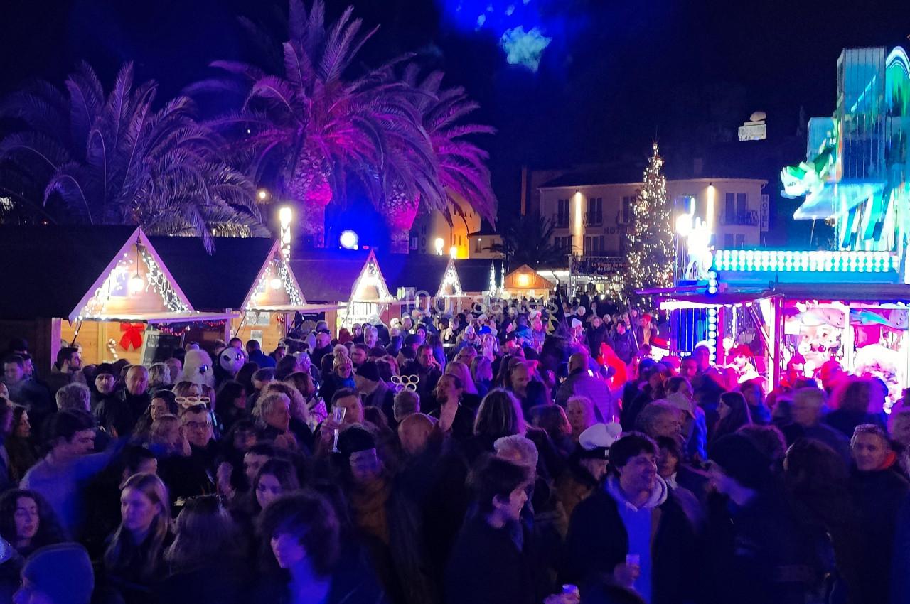 Foule rassemblée dans un marché de Noël nocturne éclairé par des lumières colorées, avec des stands et un sapin décoré.