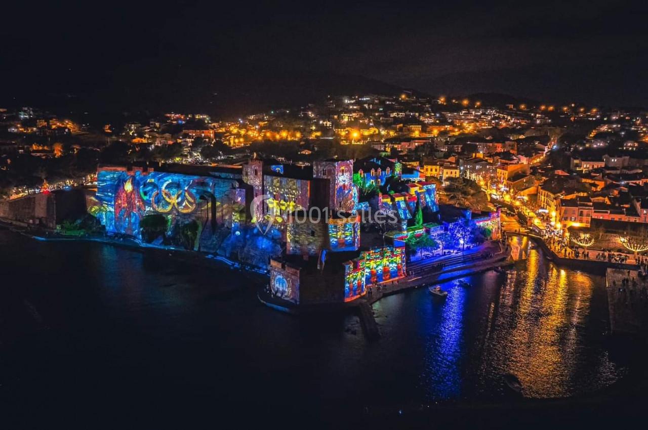 Vue nocturne d'une ville côtière illuminée avec des projections colorées sur un château et des lumières dans les rues.
