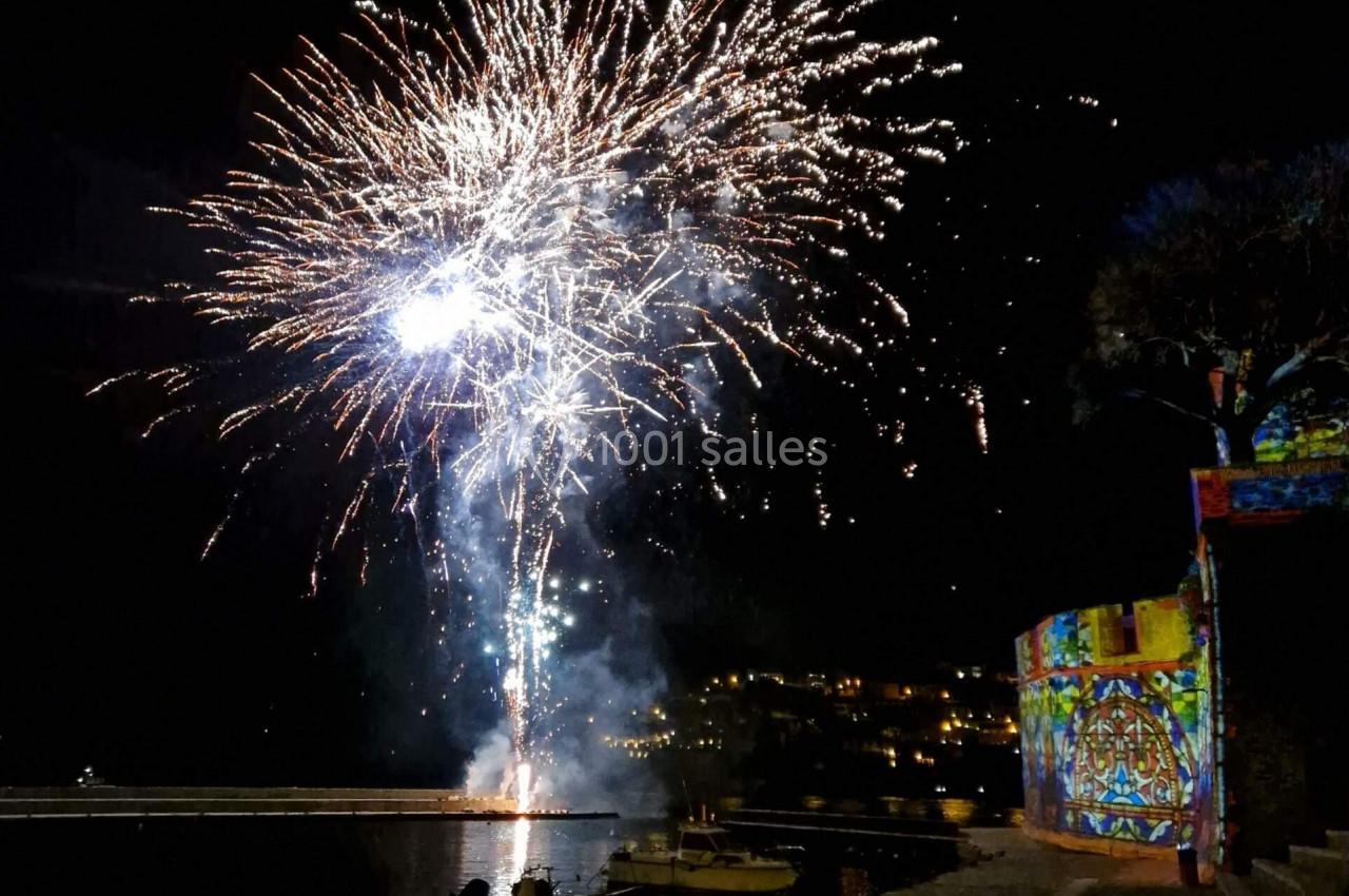 Feu d'artifice illuminant le ciel nocturne au-dessus d'un quai avec des barques et une façade colorée éclairée.