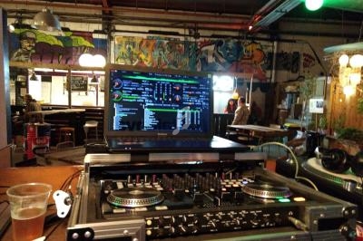 Table de mixage avec ordinateur affichant un logiciel de DJ, dans un bar décoré de graffitis colorés.