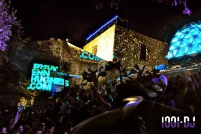 Foule dansante en soirée en plein air avec projections lumineuses sur des murs en pierre et une structure géométrique…