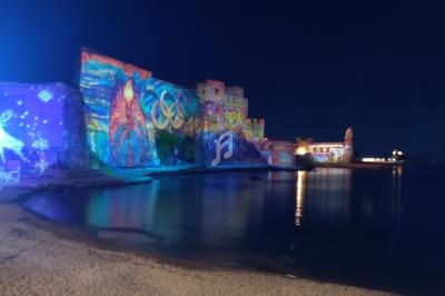 Mur illuminé avec des fresques colorées reflétées dans l'eau, près d'une plage et d'un bâtiment éclairé la nuit.