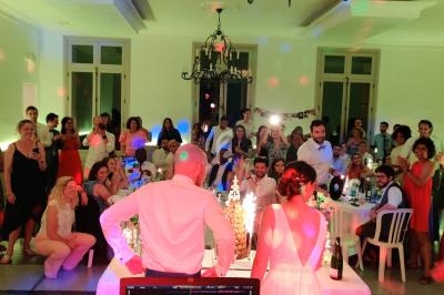 Un couple de mariés face à leurs invités dans une salle éclairée avec des lumières colorées, vue depuis une table de DJ.