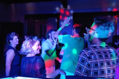 Des personnes dansent sous des lumières colorées dans une ambiance festive, avec une table de mixage visible au premier plan.