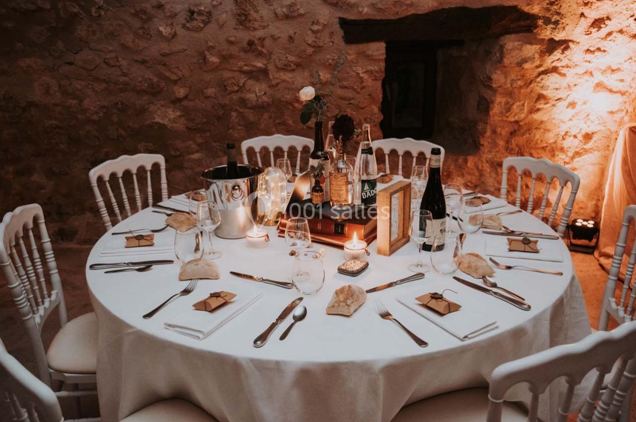 Table ronde décorée pour un dîner, avec nappes blanches, couverts, bougies et cadeaux sur chaque assiette.