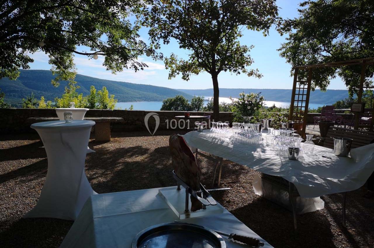 Vue d'une terrasse ombragée avec tables dressées, offrant un panorama sur un lac entouré de collines verdoyantes.