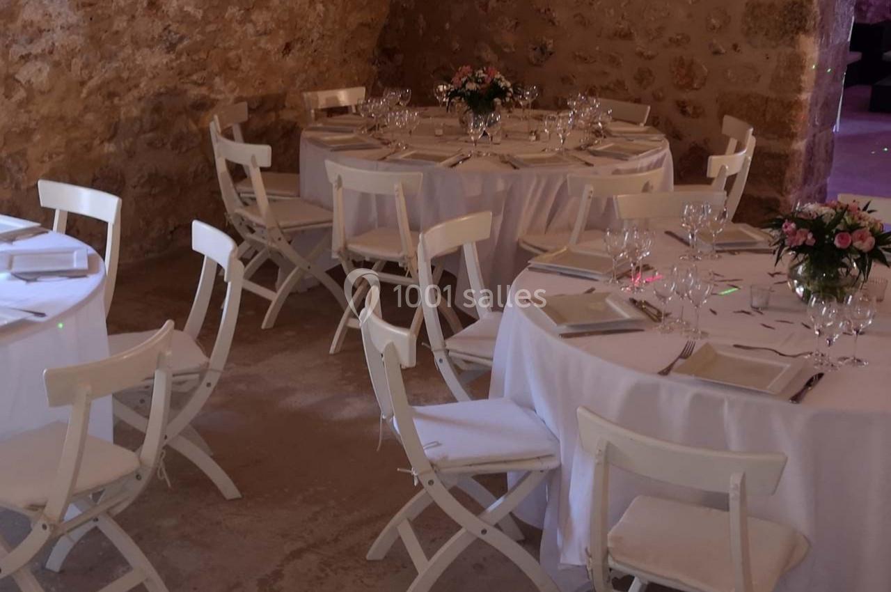 Salle en pierre aménagée avec des tables rondes dressées de nappes blanches, vaisselle et bouquets de fleurs.