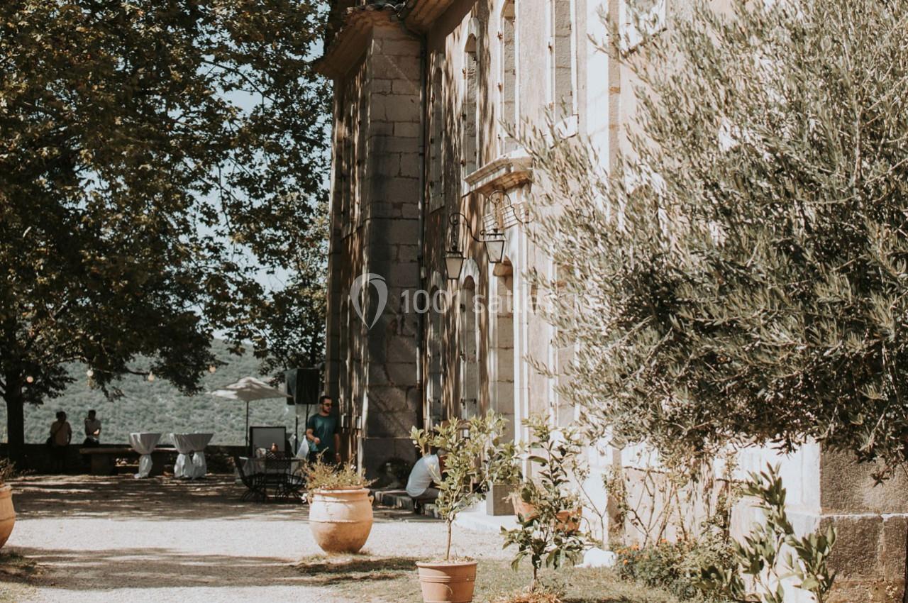 Façade en pierre d'un bâtiment ancien entouré de végétation, avec des tables installées en extérieur.
