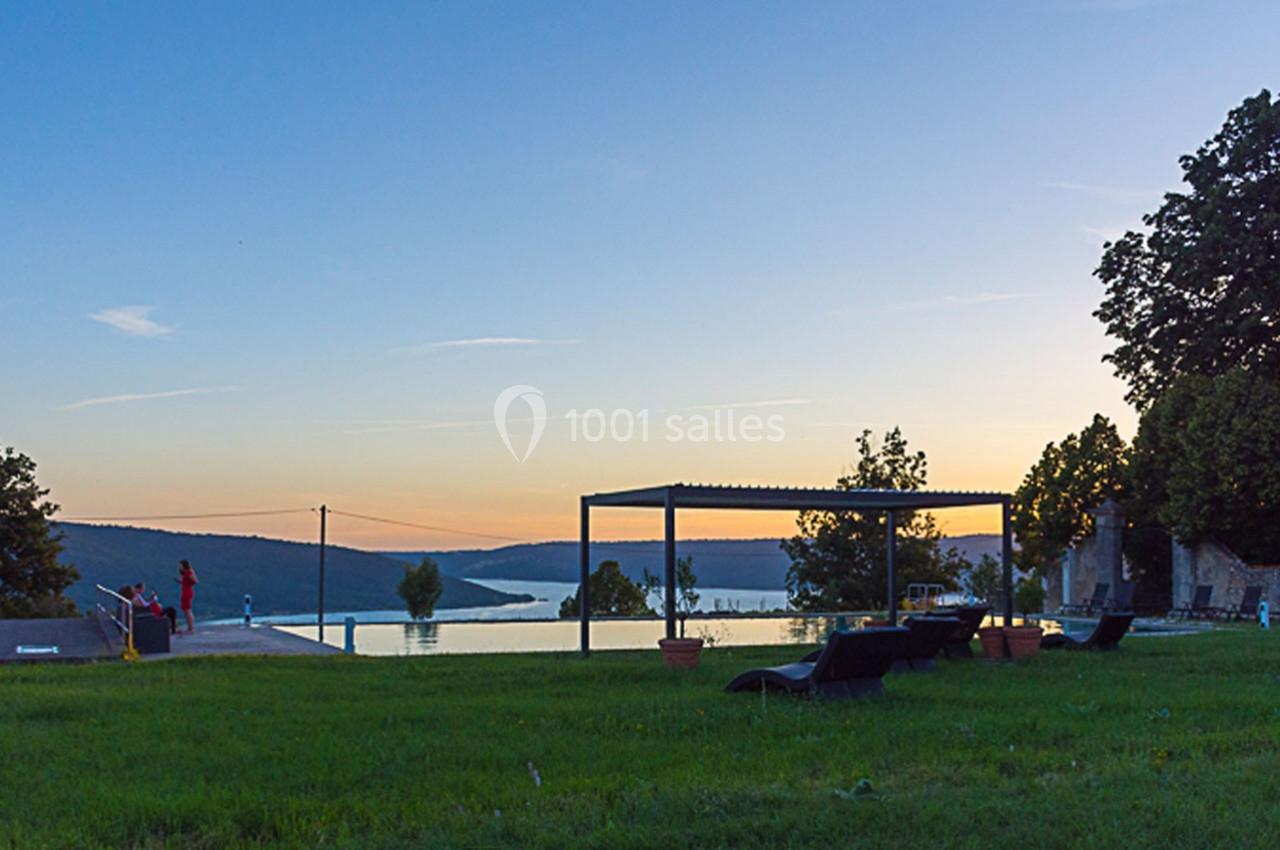 Vue d'une piscine à débordement entourée de chaises longues, avec un paysage de lac et collines au coucher du soleil.