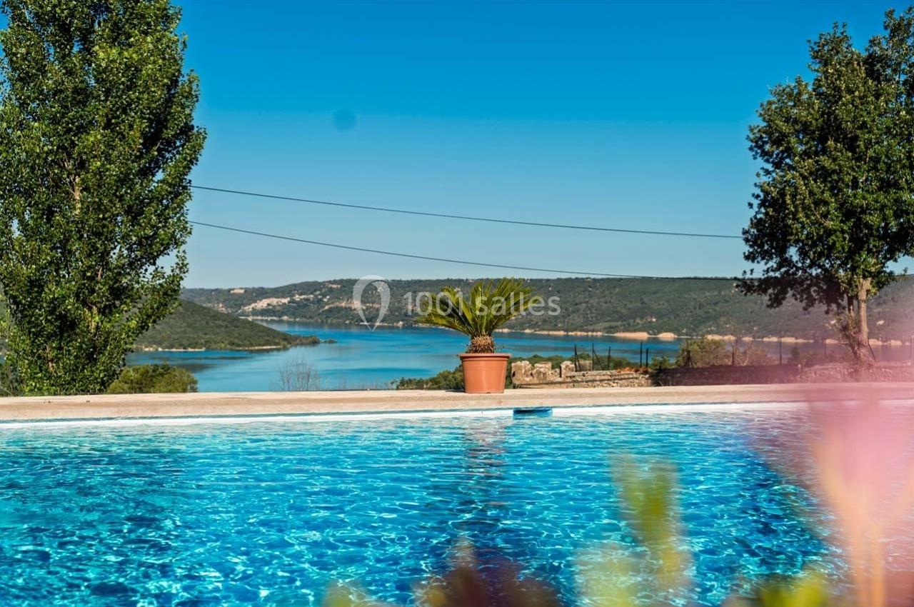 Vue d'une piscine extérieure avec un pot de palmier, des collines et un lac en arrière-plan sous un ciel bleu.