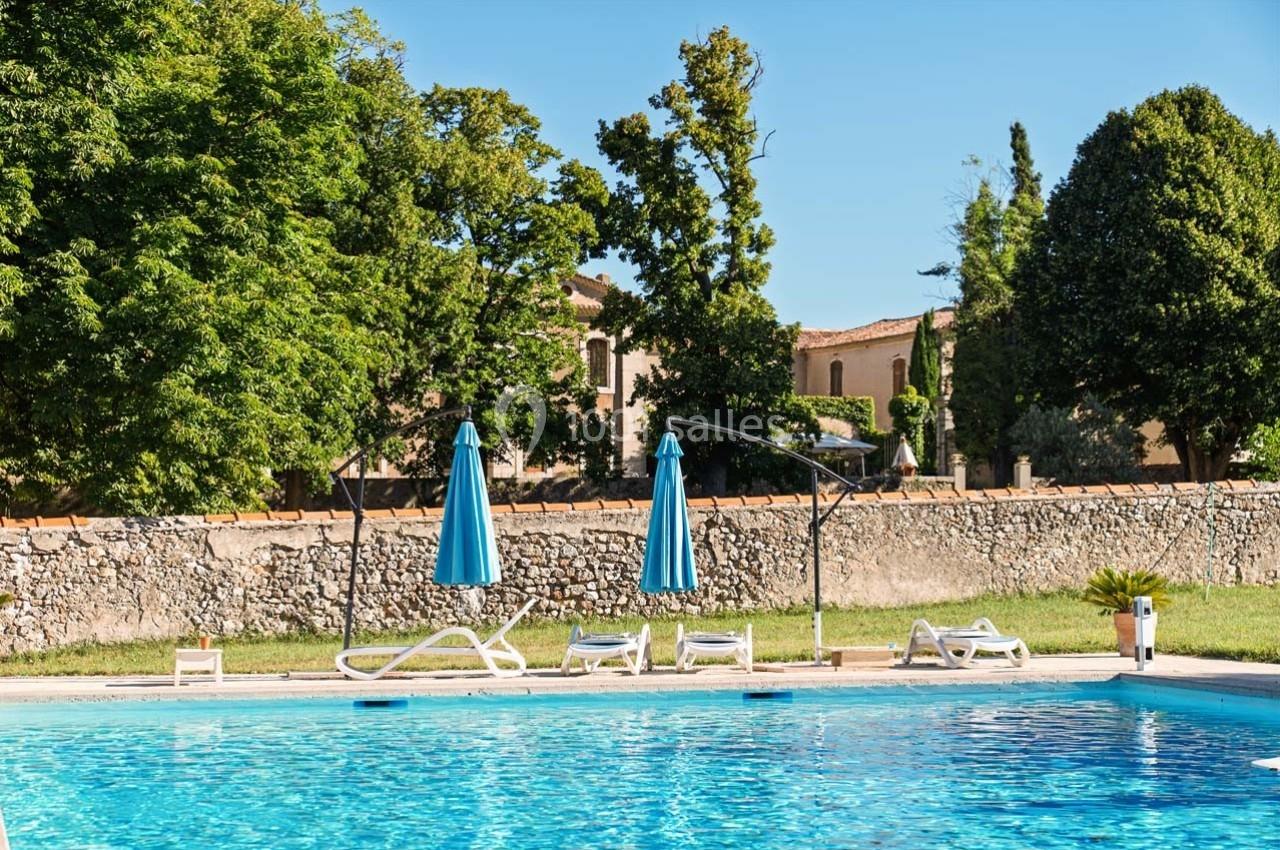 Piscine extérieure entourée de chaises longues, parasols bleus et arbres, avec un bâtiment en arrière-plan.
