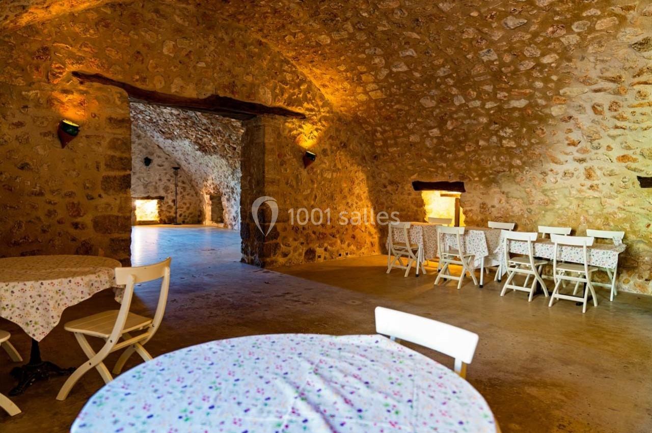Salle voûtée en pierre avec tables et chaises blanches, éclairée par des appliques murales et une lumière naturelle.