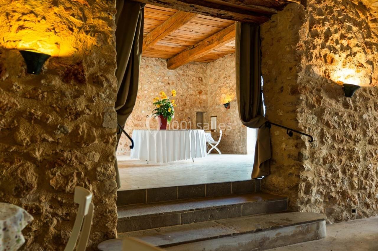 Salle en pierre avec plafond en bois, table blanche décorée de fleurs jaunes et rideaux encadrant l'entrée.