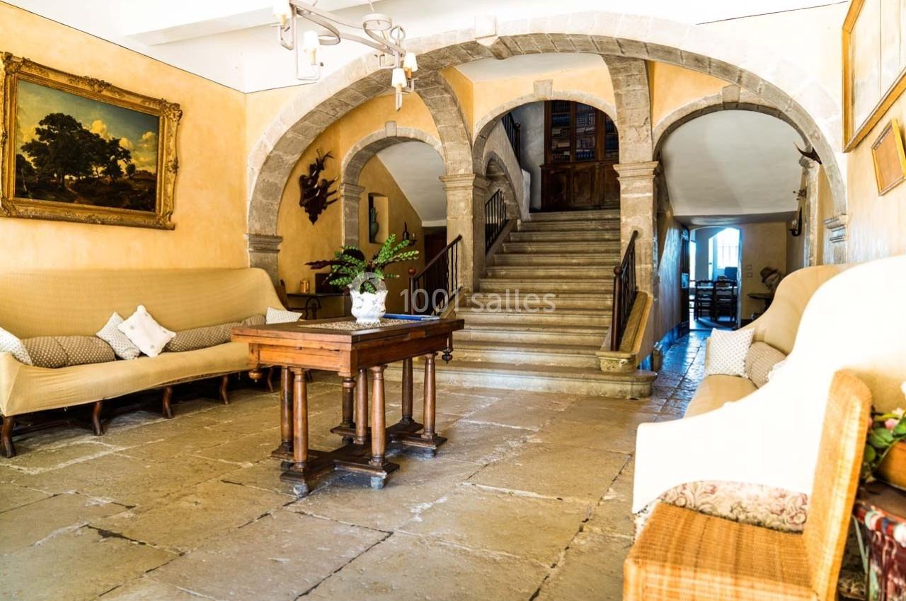 Salon lumineux avec canapés, table en bois, plante en pot, arches en pierre et escalier menant à l'étage.