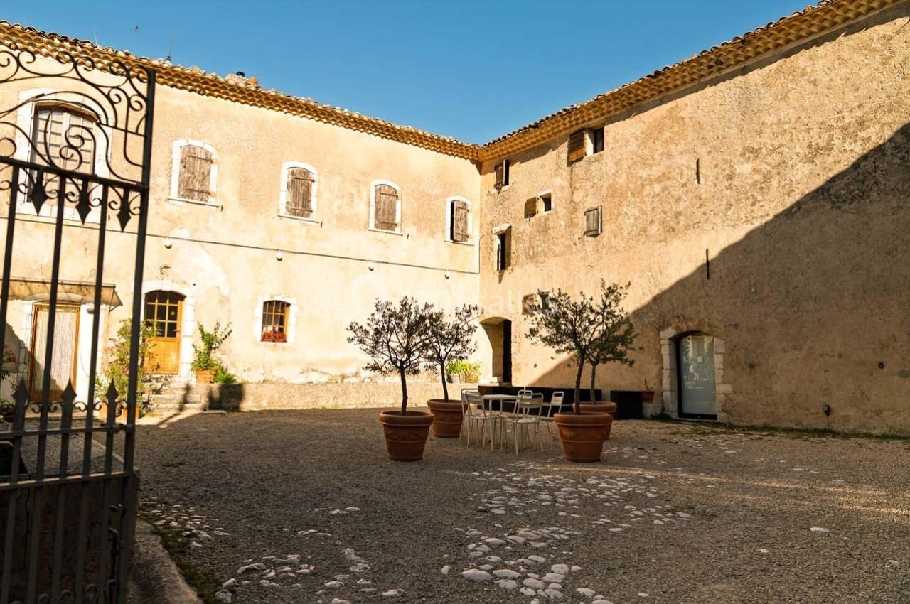 Cour intérieure ensoleillée avec des murs en pierre, des pots d'oliviers et une table entourée de chaises blanches.