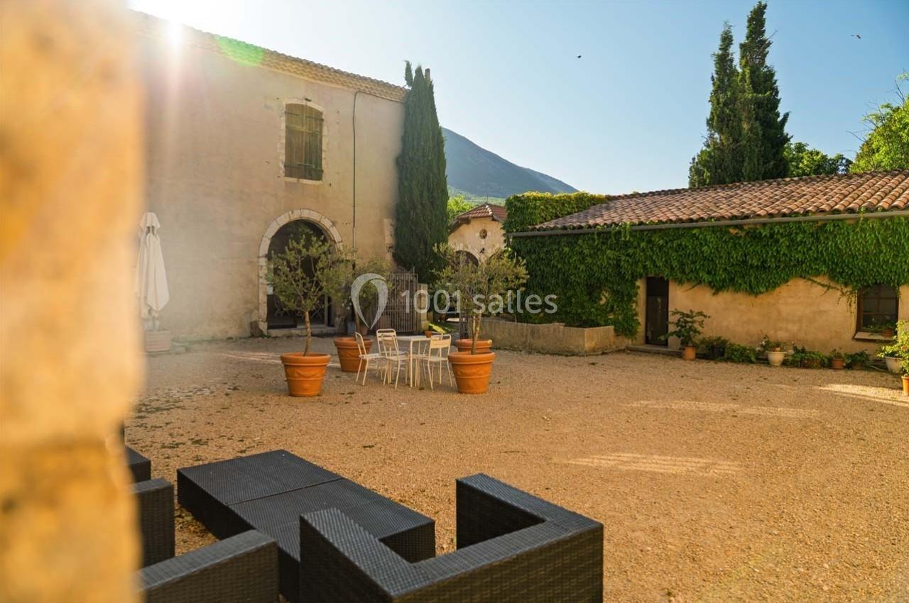 Cour ensoleillée avec mobilier de jardin, pots d'oliviers et bâtiments en pierre recouverts de végétation.