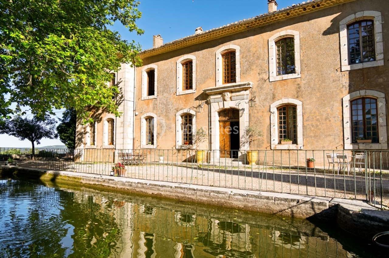 Façade d'une maison en pierre avec des volets en bois, bordée par un bassin et entourée de végétation.