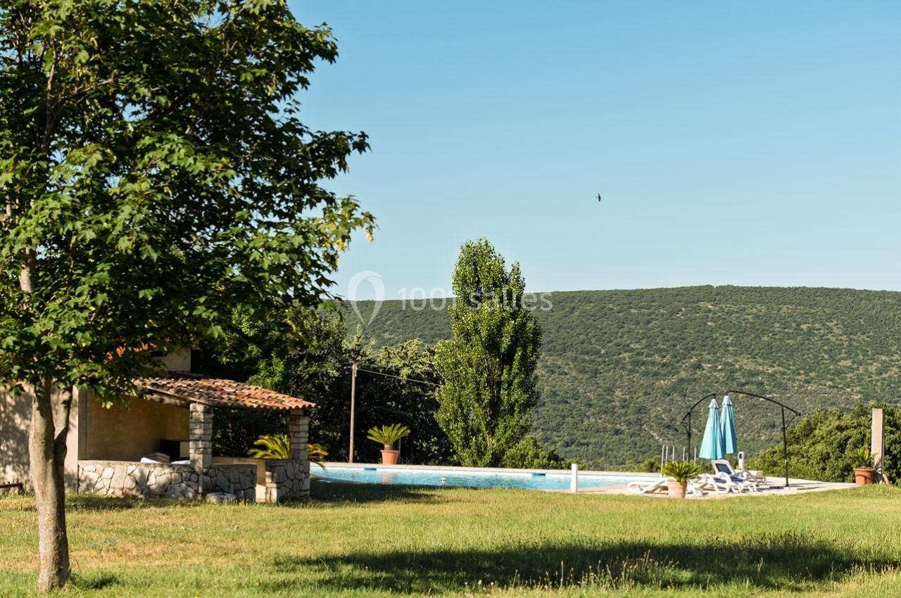Jardin avec pelouse, piscine entourée de chaises longues et parasols, vue sur des collines verdoyantes.