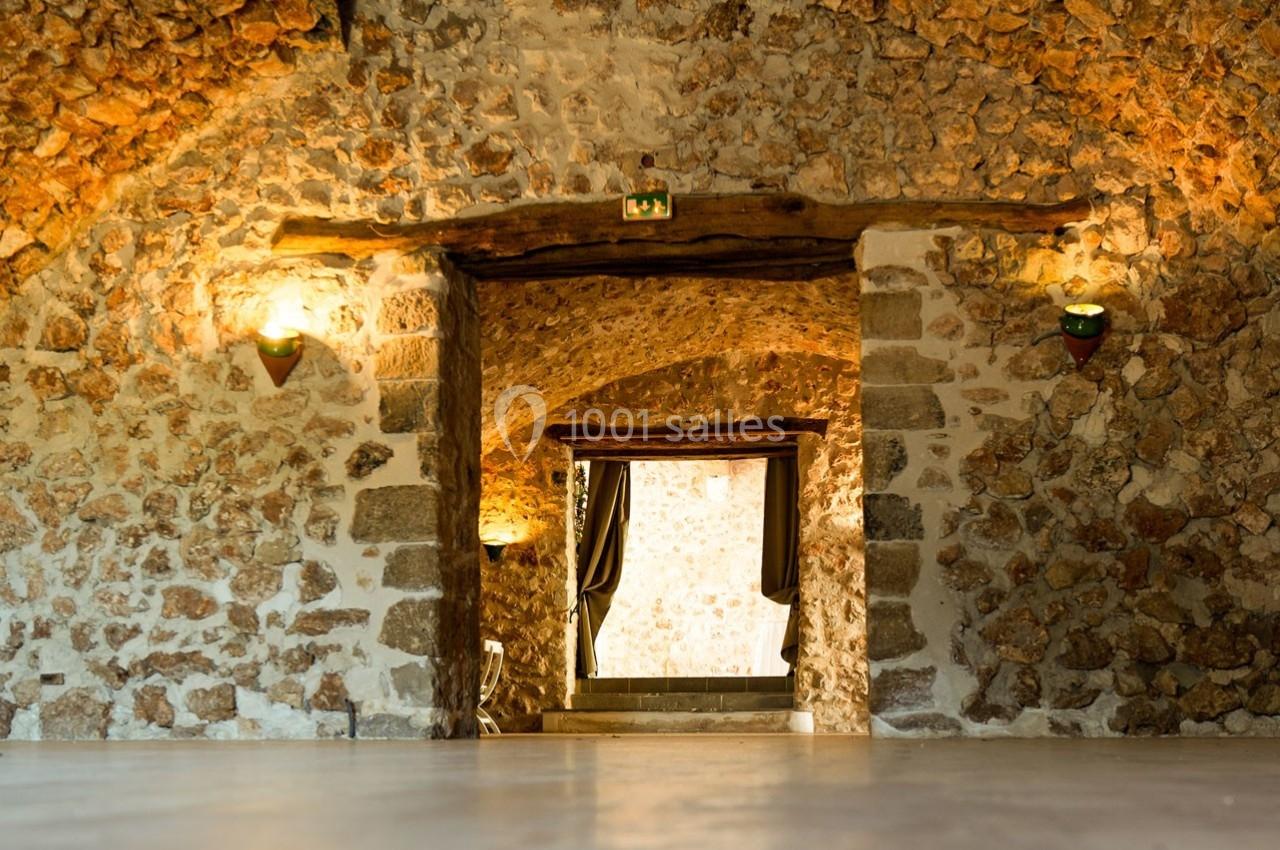 Salle voûtée en pierre avec éclairage mural, ouvrant sur une porte encadrée de rideaux et une lumière naturelle.