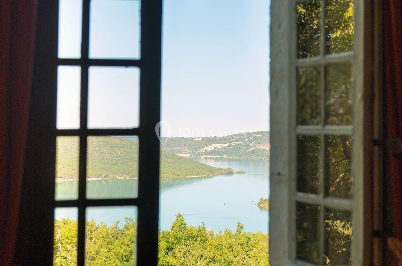 Vue sur un lac entouré de collines verdoyantes à travers une fenêtre ouverte avec des volets en bois.